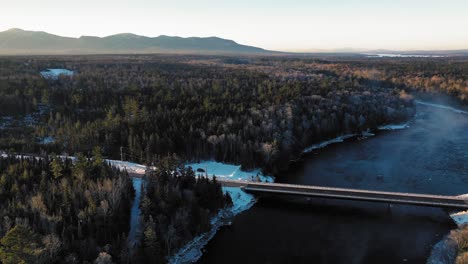 Luftumlauf-Um-Eine-Brücke,-Die-Einen-Winterlichen-Fluss-überquert-Und-Einen-Bergrücken-Enthüllt,-Der-In-Der-Frühen-Morgensonne-Erleuchtet-Ist