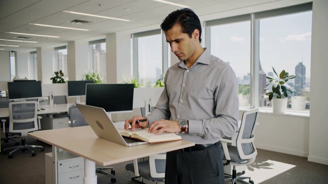 Hombre comenzando a trabajar, consultando un cuaderno y escribiendo en un portátil en un escritorio de pie en la oficina, con camisa gris.