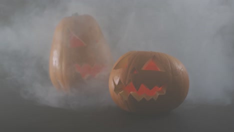 Smoke-effect-over-two-scary-face-carved-halloween-pumpkin-against-grey-background
