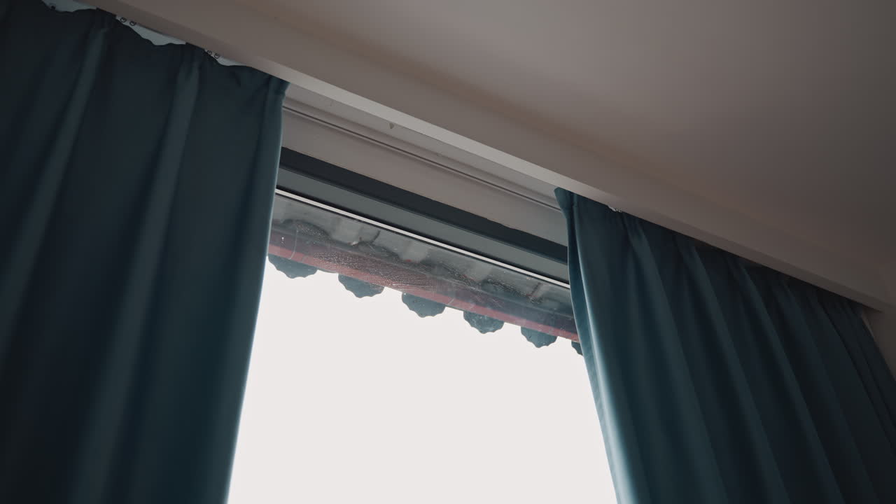 Luce mattutina attraverso le tende della finestra dell'hotel, tenda color verde acqua parzialmente aperta che rivela una tenda da sole piastrellata e un cielo pallido, modanatura del soffitto morbida, delicata luce calda, atmosfera accogliente della suite, suggerendo una scena di risveglio dell'ospite.