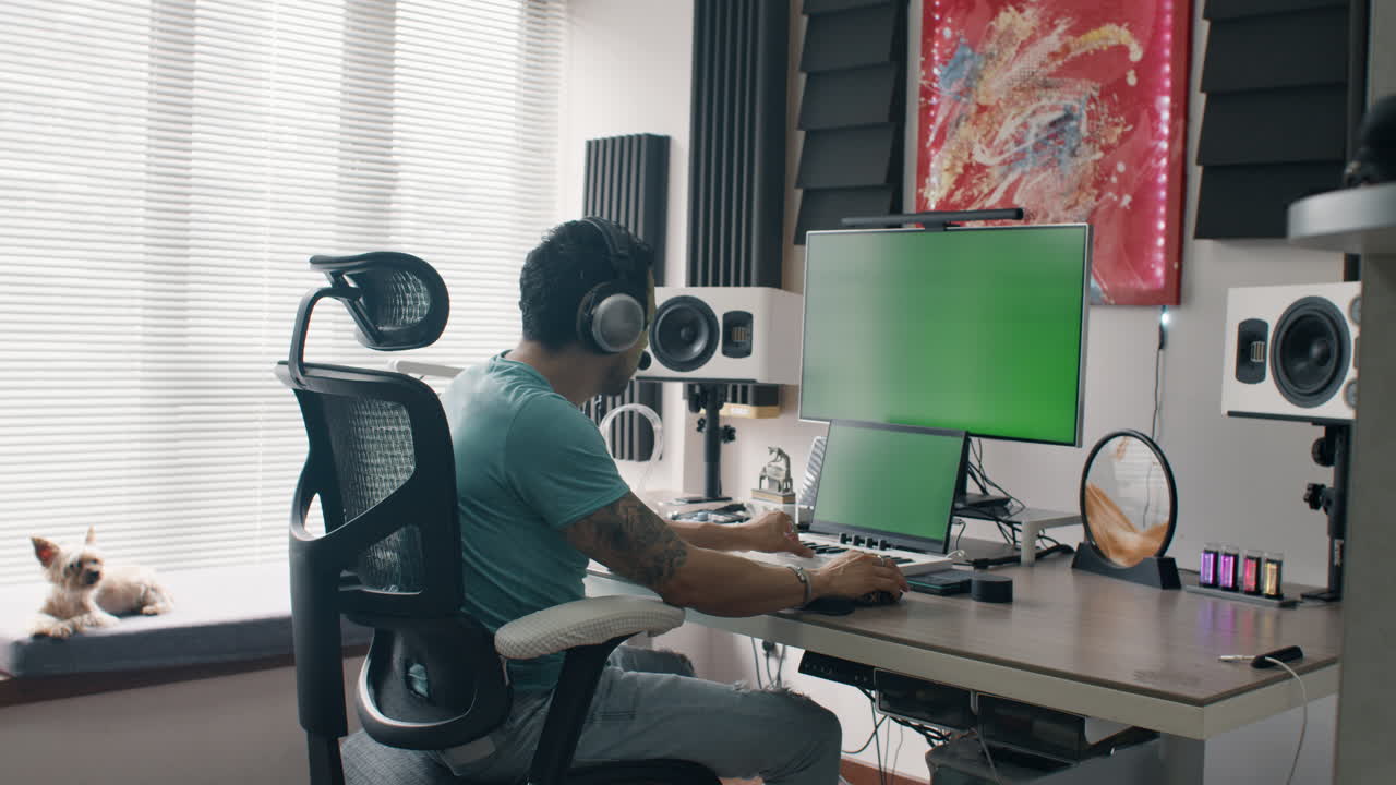 Person working in a home music studio with headphones, dog resting by the window, green screen