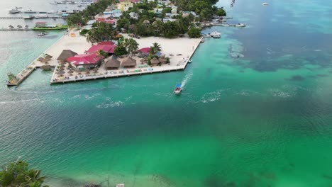 vista de avión no tripulado en belice volando sobre el mar verde y azul del caribe, un cay de arena blanca con palmeras y restaurantes con un barco cruzando un canal en un día nublado