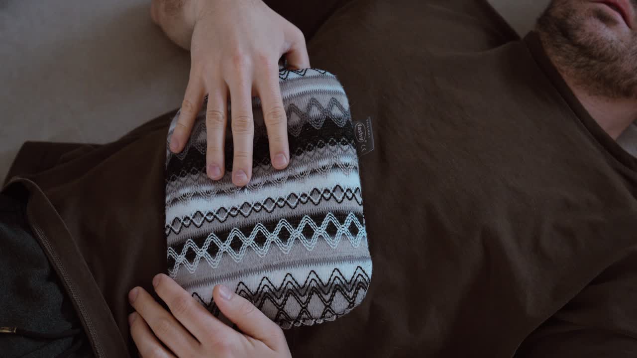 Primo piano di un uomo sdraiato con una borsa dell'acqua calda posizionata sullo stomaco. Concetto di dolore addominale, crampi, mal di stomaco, rimedio casalingo e trattamento di auto-cura.