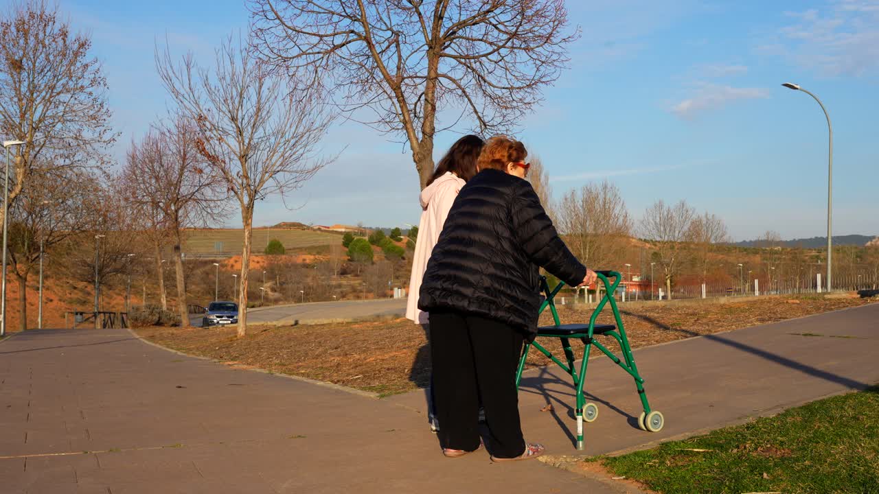 Elderly woman walking with walker assisted by young woman in park