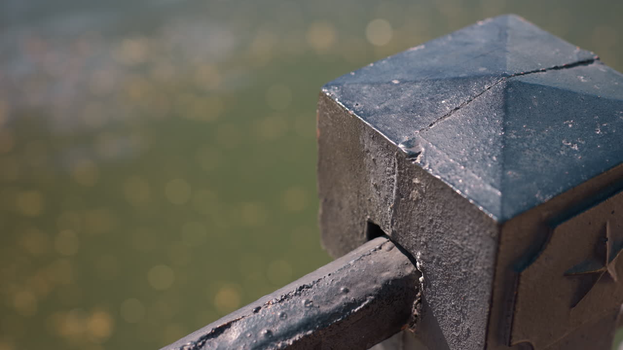 stone railing post with blurred pond background, close-up capture of weathered concrete cap and metal rod, sharp texture and chipped patina, soft bokeh from distant water, warm sunlight highlights