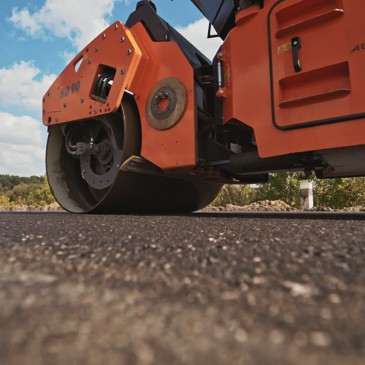 Orange roller machine pressing hot asphalt on the road in summer. Heavy compactor machine making new asphalt on the background of blue sky and green trees. Road resurfacing.