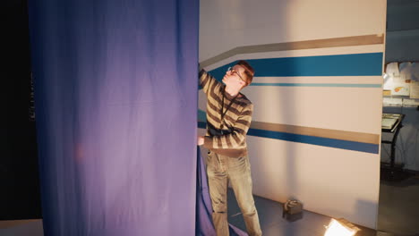 male stagehand pulls blue curtain aside in television studio during set preparation, floor spotlight illuminates equipment, ladder and cables visible backstage ready for live broadcast production