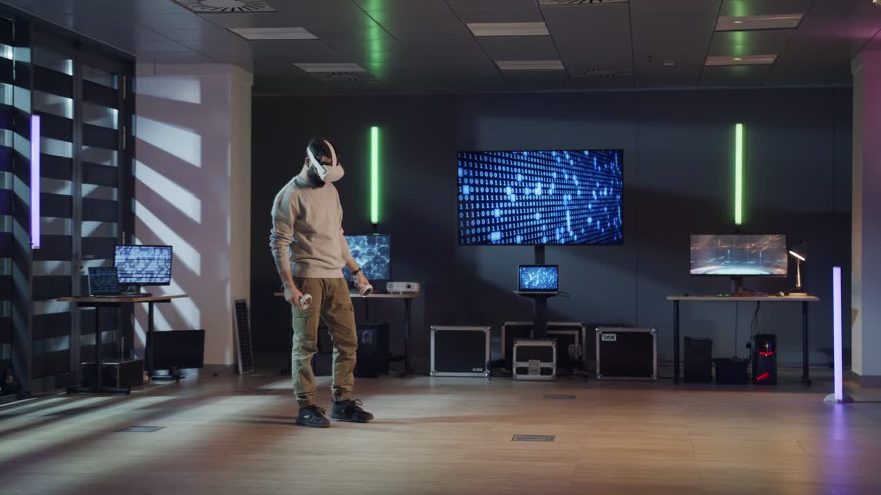 Wide shot of a man in a VR headset testing a virtual reality application in a modern, high-tech office. The room is filled with computers and neon lights, suggesting a VR development or gaming lab.