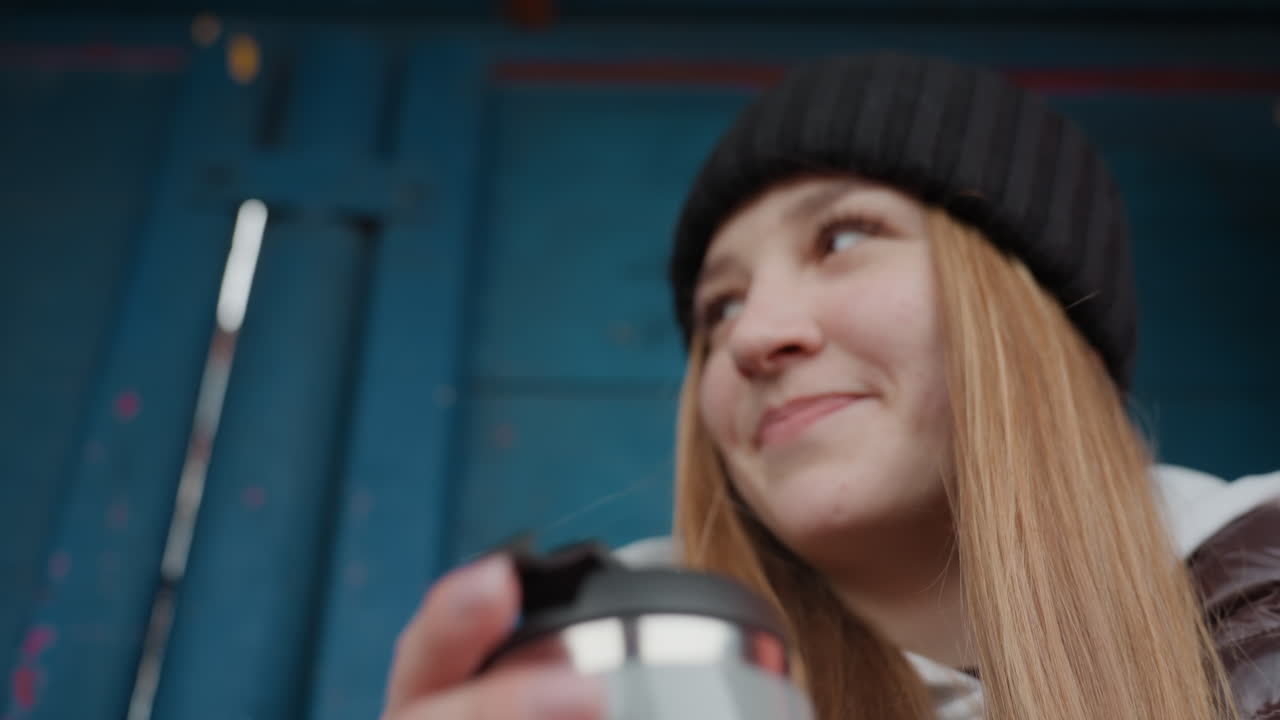 una mujer blanca saboreando un café caliente en una taza de cerámica en un refugio azul, con pelo largo y gorro de punto, chaqueta marrón y sudadera con capucha, las manos sosteniendo la taza, pausa relajada en la calle con un ambiente acogedor y tonos suaves