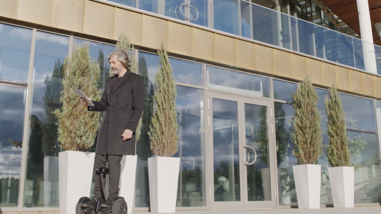 Businessman on self-balancing scooter using tablet outside modern office building