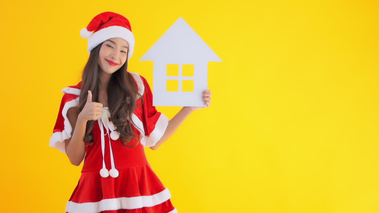 A young woman dress as Santa's helper hold up a symbol for a house then gives the thumbs up and smiles. Copy space