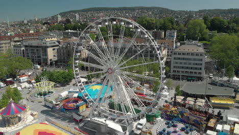 Hermosa-Grúa-Giratoria-Con-Drones-Aéreos-Tomada-De-La-Rueda-De-La-Fortuna-Del-Parque-De-Diversiones-Con-La-Ciudad-De-Zúrich,-Suiza-En-El-Fondo-Durante-El-Zürichfest