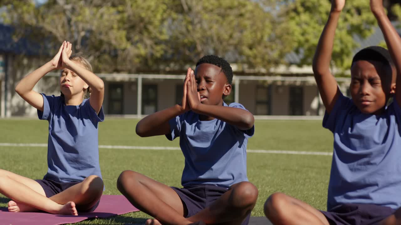 Diversos estudantes adolescentes levantando os braços e unindo as palmas das mãos em exercícios de respiração guiada em colchonetes de ioga em um campo de esportes