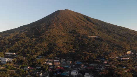 forwards flying aerial drone over beautiful single hill on green landscape with small village at bottom