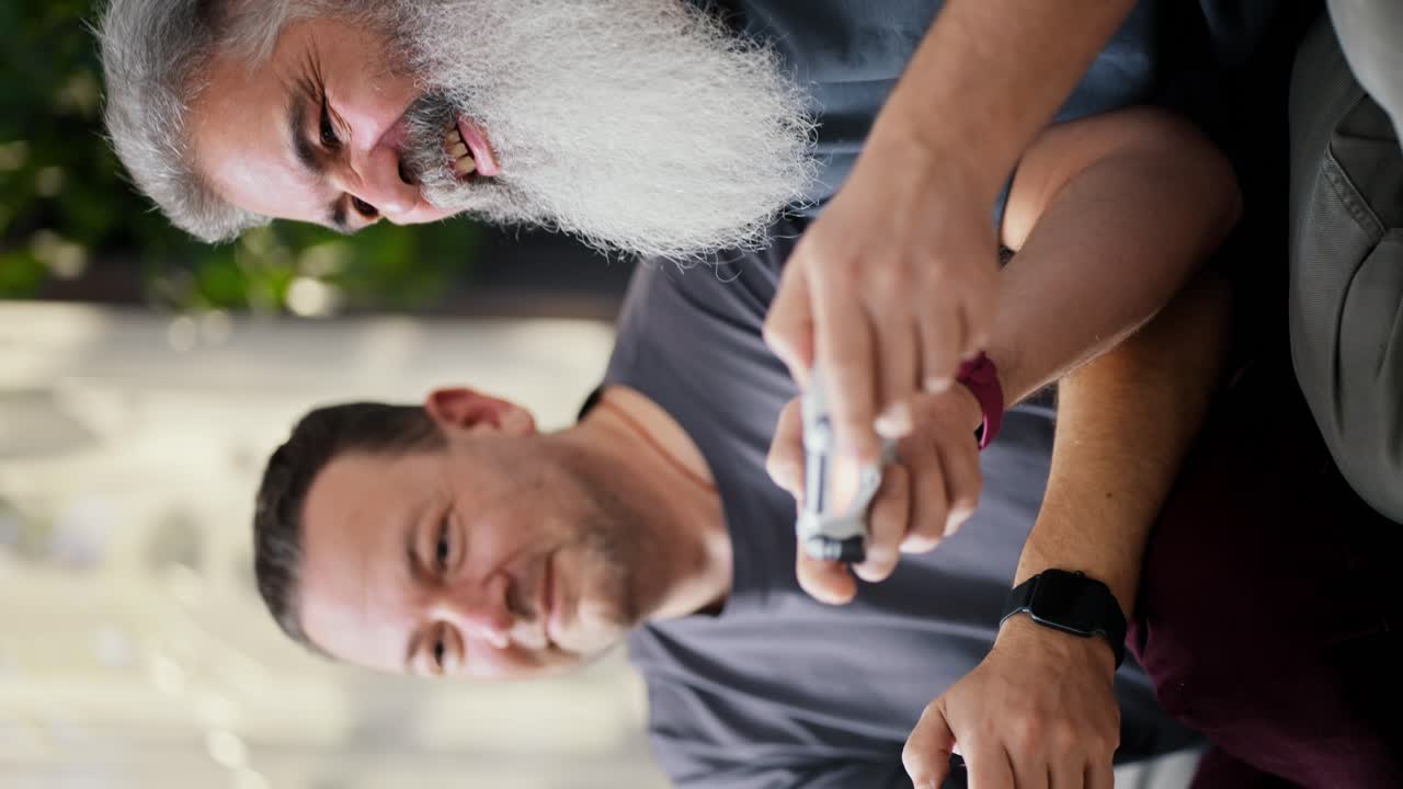 Vertical video of a happy elderly man with gray hair and a lush gray beard playing video games using a joystick together with his brunette boyfriend with stubble in a gray T-shirt on the sofa in a modern apartment LGBT couple