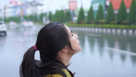 beautiful carefree asain woman stands on city roadside looking up to sky check for raining, season changes or global warming concept, walking outside in rain, waterproof make up, getting fresh in rain