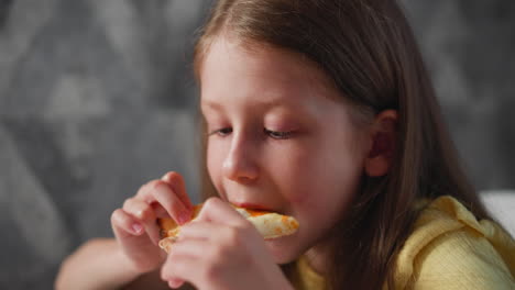 close up of cute girl in yellow shirt eating cheesy pizza while seated indoors against textured wall background enjoying meal with focus on expression and hands holding delicious slice of pizza