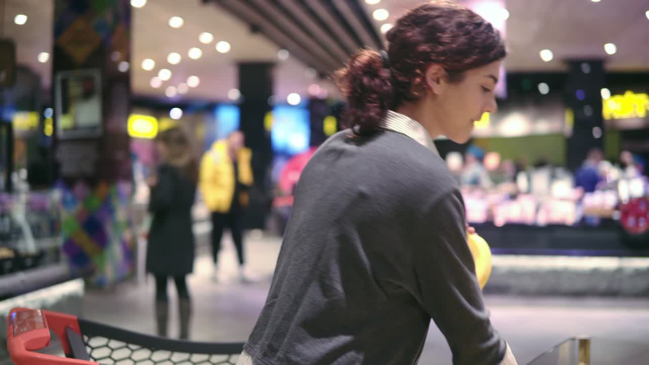 Young beautiful brunette girl in her 20's picking out bananas and putting them into shopping cart at the fruit and vegetable aisle in a grocery store