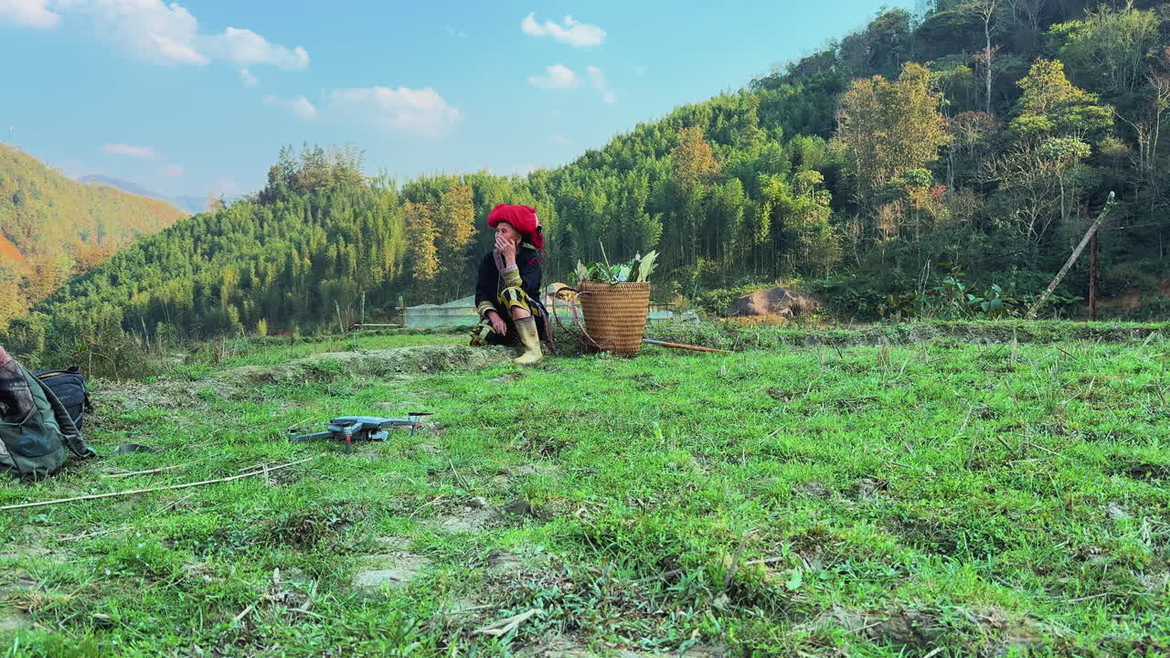 A Black Dao woman in traditional dress sits beside her harvest basket in a green field. She looks back at the end.