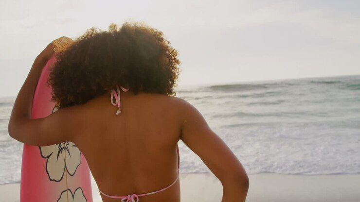 Vista posteriore di surfista afroamericana in piedi con la tavola da surf sulla spiaggia 4K