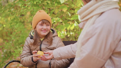 sad girl in brown jacket and orange beanie sits with head down on park bench as caring mother offers fruit with gentle hand gesture during quiet autumn moment surrounded by green and golden foliage