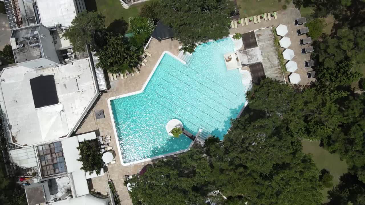 Aerial rotating view revealing expansive swimming pool nestled among lush trees, surrounding resort buildings under bright summer sunlight