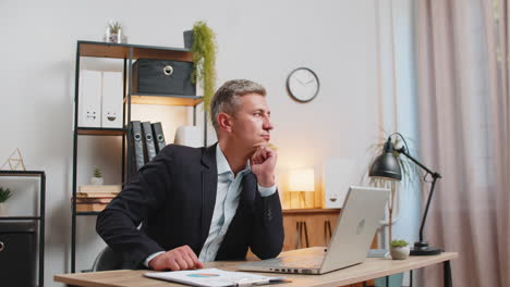 a man in a suit sits at a desk with a laptop, thinking in his office