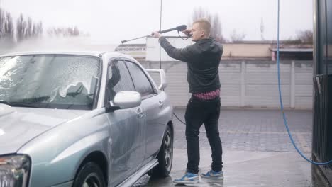 Young-attractive-stylish-man-is-carefully-washing-his-silver-sportcar-with-water-jet-on-self-service-carwash.-Slowmotion-shot