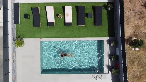 drone flies over an outdoor pool in bird's eye view - woman in swimming costume swims from one side to the other - villa in greece crete with palm trees