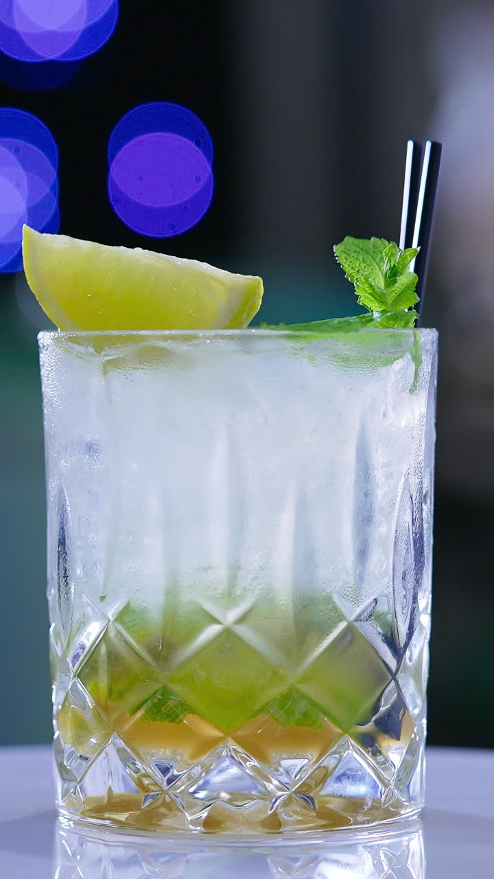 Misted glass full of beverage with two short black straws in it. Rotating glass with lemon and mint leaves on top. Close up. Blurred backdrop. Vertical video