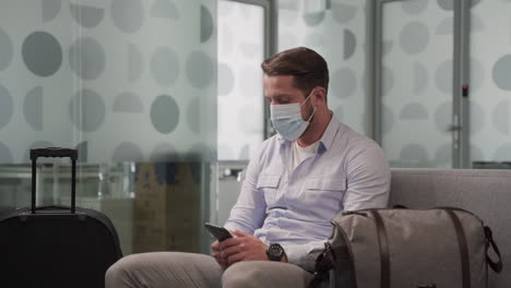 a young man wearing a mask consults his cell phone while waiting for his flight to depart in an airport lounge.