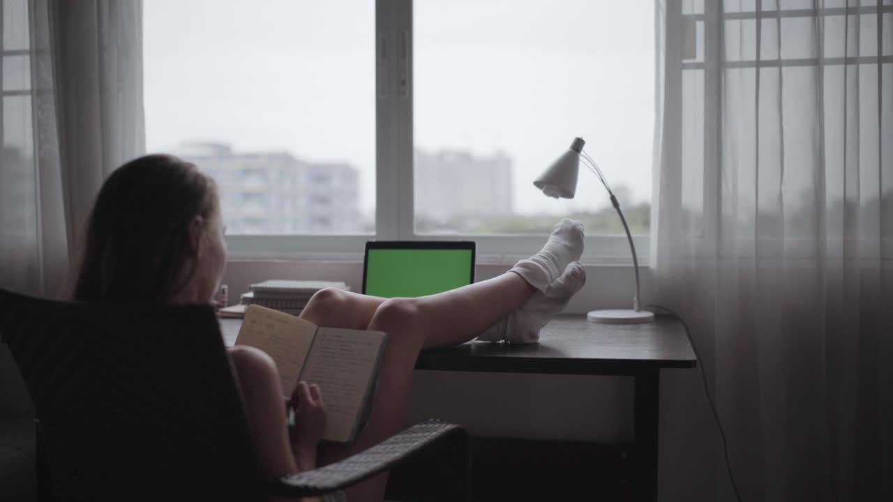 작업 테이블에 발을 올려놓고 앉아 메모장에 글을 쓰고 노트북의 녹색 화면을 보는 젊은 여성