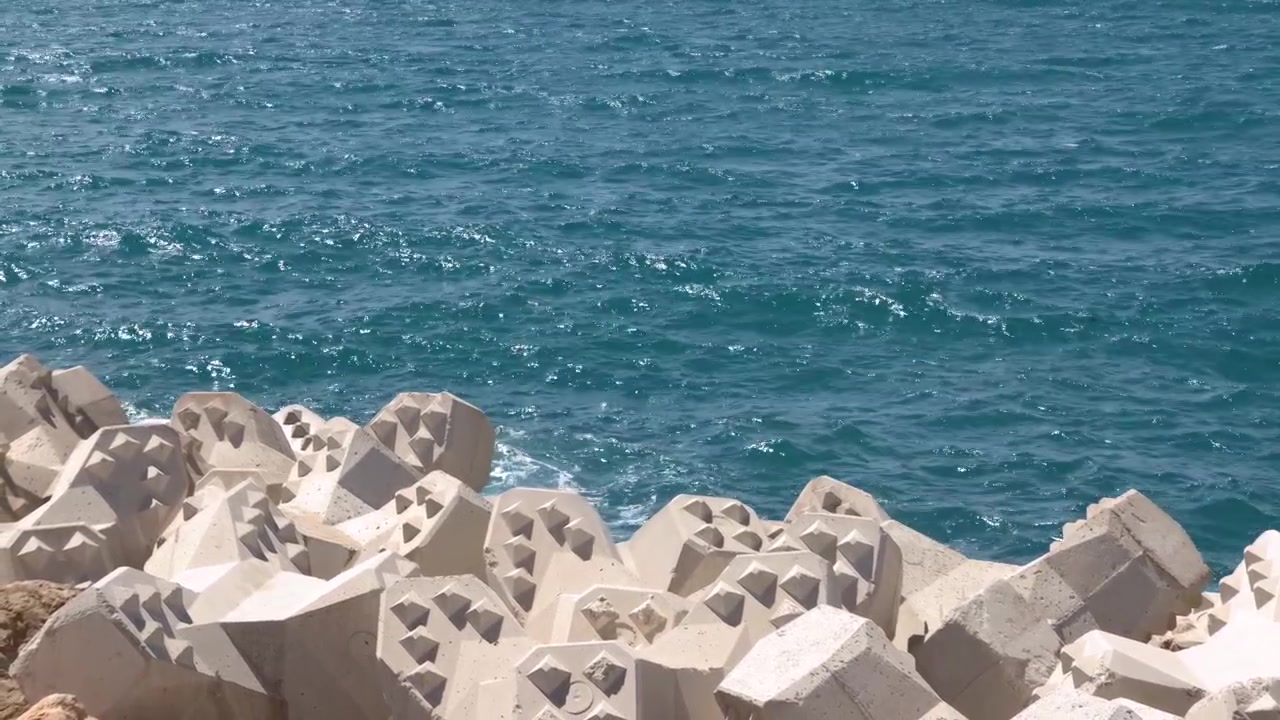 Static wide shot of blue ocean waves hitting a concrete tetrapod seawall under bright sunlight.
