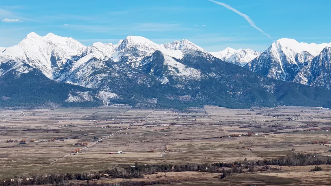 Una vasta vista panoramica delle Montagne Rocciose innevate che si ergono su una valle rurale vicino a Whitefish, Montana, con campi aperti e edifici agricoli sparsi in primo piano.