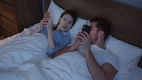 joven hombre y mujer caucásicos sonrientes acostados en la cama por la noche y jugando en los teléfonos inteligentes, luego se vuelven el uno al otro