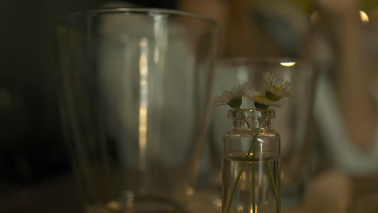 Statische close-up van enkele kleine bloemetjes in een vaas naast een glas op een tafel