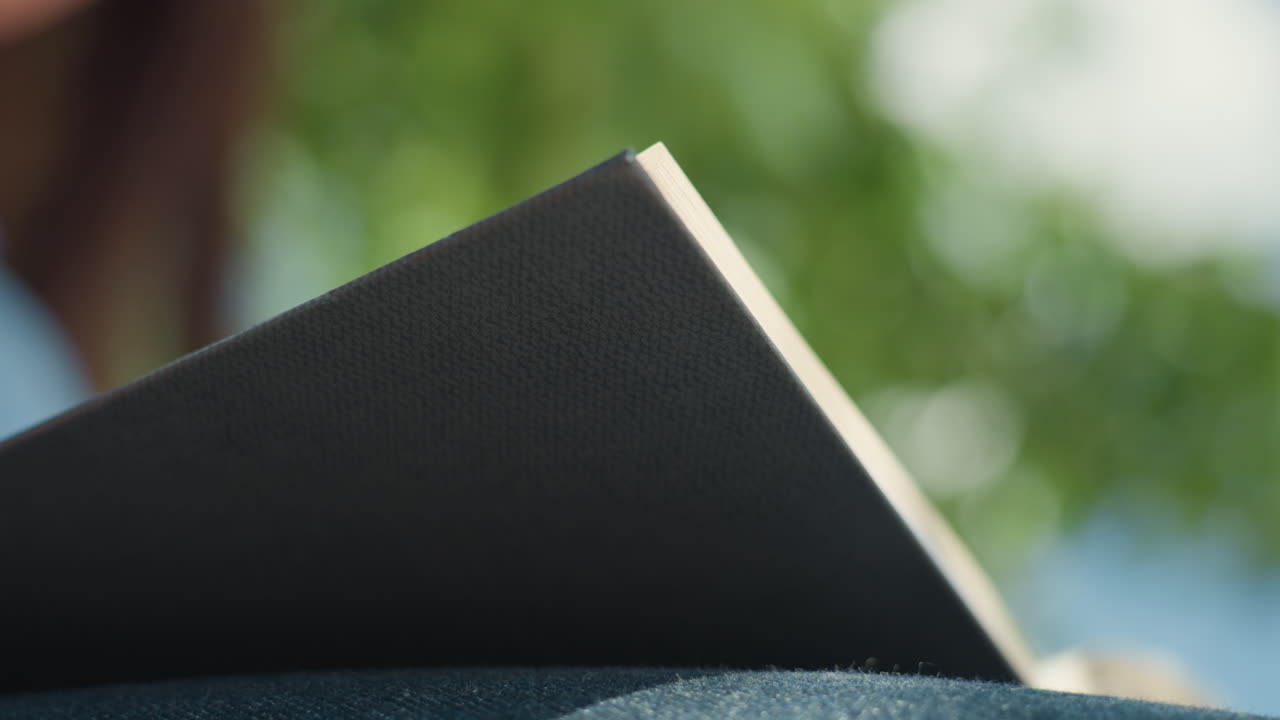 Primer plano de una mano tocando la cubierta texturizada de un libro, contacto suave con la punta de los dedos con un libro de tapa dura en un entorno natural, escena detallada de una mano acariciando un libro texturizado bajo la luz del sol entre el follaje.