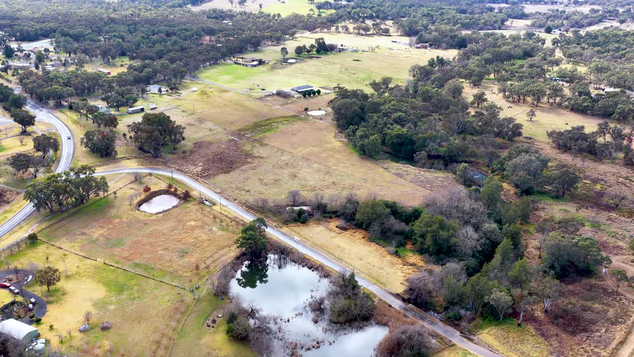 Drone footage glides above a rural area with ponds, roads, scattered houses, and trees under soft daylight, capturing a peaceful countryside environment