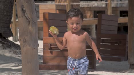 niño latino joven caminando en la playa en un día soleado usando sus pantalones cortos de traje de baño y llevando un coco