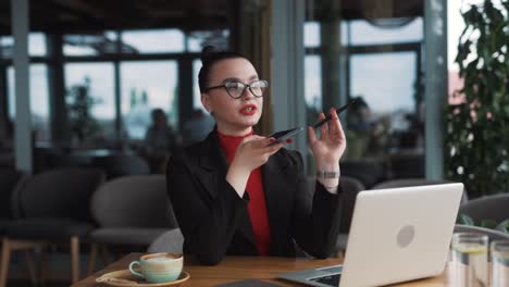 Eine-Schöne-Junge-Frau-Mit-Brille-Und-Zahnspange,-Eine-Freiberuflerin,-Sitzt-In-Einem-Stilvollen-Restaurant-In-Professioneller-Kleidung-Und-Nimmt-Eine-Sprachnachricht-Auf-Ihrem-Smartphone-Auf,-Während-Sie-An-Ihrem-Laptop-Arbeitet