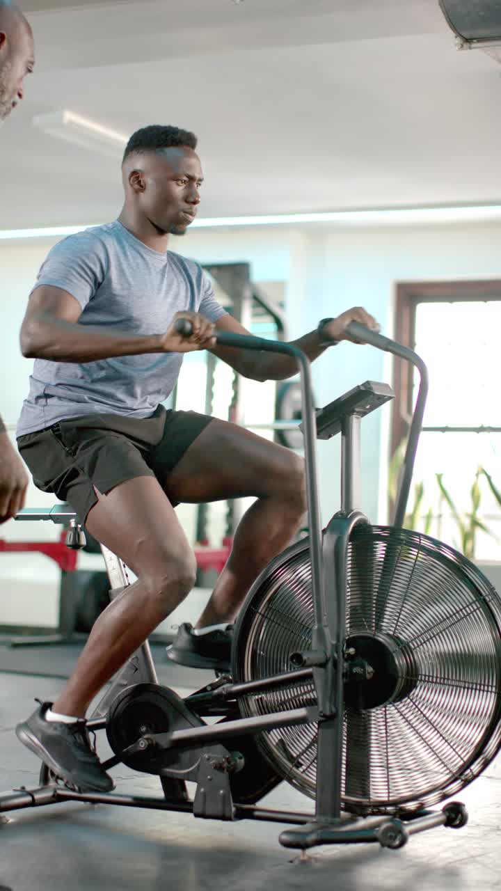 Video verticale: Allenatore che arriva e corregge la forma mentre un uomo afroamericano pedala su una airbike in palestra.