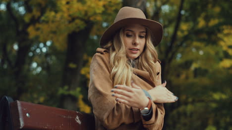 joven rubia caucásica sintiendo frío y calentándose mientras se sienta en el banco y bebe café para ir al parque en otoño