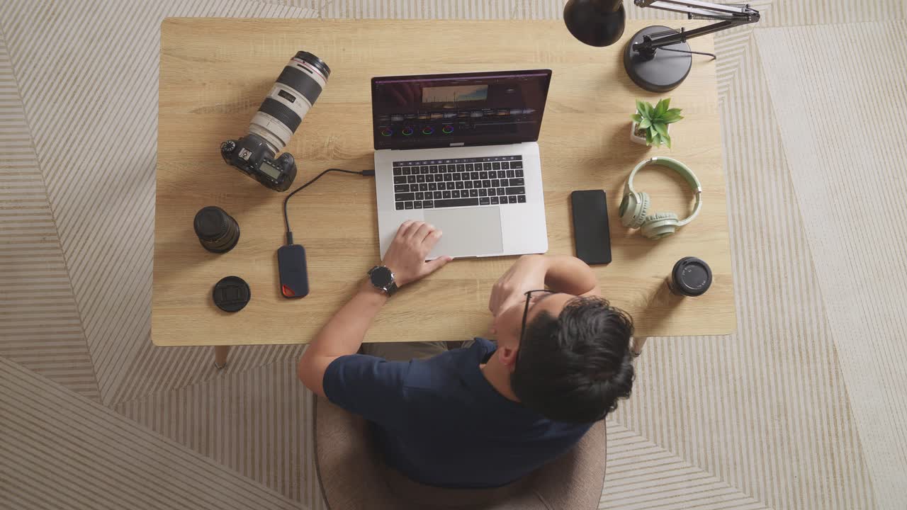 Top View Of Asian Male Color Grading About The Footage Then Raising Index Finger While Sitting In The Workspace Using A Laptop Next To The Camera Editing The Video At Home