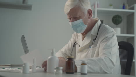 a grey haired, middle aged doctor in a facemask writes medical results from his notebook to his laptop