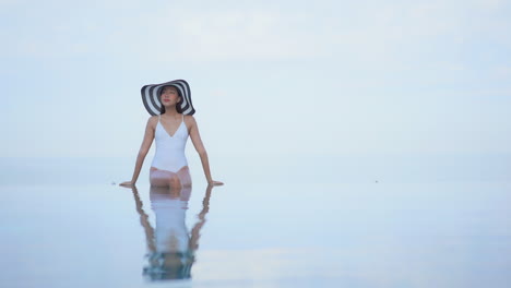 Glamorous-asian-female-model-in-swimsuit-and-summer-hat-sitting-on-infinity-pool-border