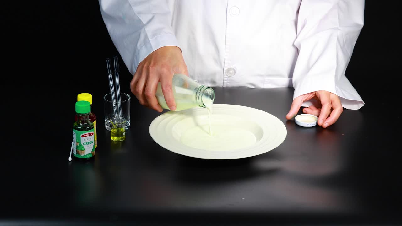 실험실 코트를 입은 사람이 통제된 환경에서 다채로운 액체로 둘러싸인 접시에 우유를 부어줍니다.
