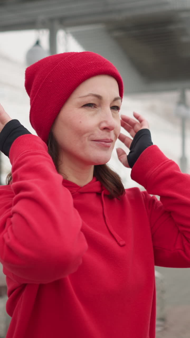 Middle-aged woman in red hoodie adjusts her beanie outdoors during winter, warm smile on her face with blurred figures in background and urban structures visible under snowy conditions
