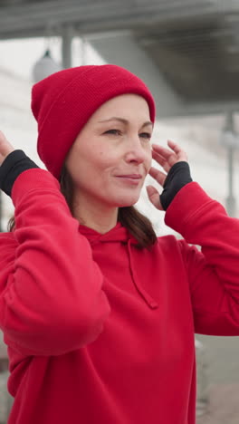 mujer de mediana edad con capucha roja ajusta su gorra al aire libre durante el invierno, sonrisa cálida en su rostro con figuras borrosas en el fondo y estructuras urbanas visibles bajo condiciones de nieve