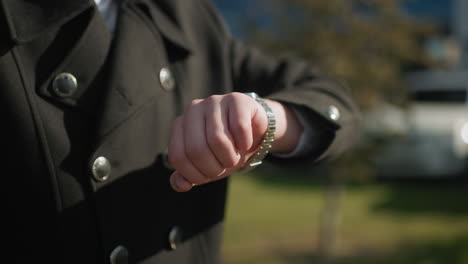 close-up of elegant black coat featuring shiny metallic buttons in outdoor urban setting as individual checks wristwatch, background includes modern buildings, parked cars, and bright lighting