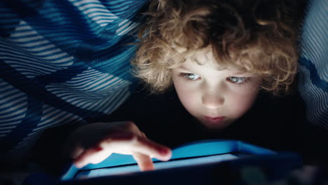 niño feliz usando una tableta digital bajo una manta disfrutando de aprender en la tecnología de pantalla táctil jugando juegos divirtiéndose antes de acostarse